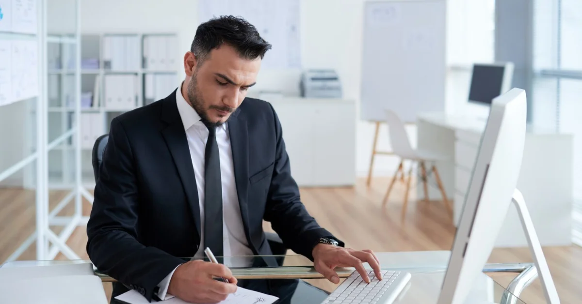 man in business suit sitting in office, working on computer and writing on a paper